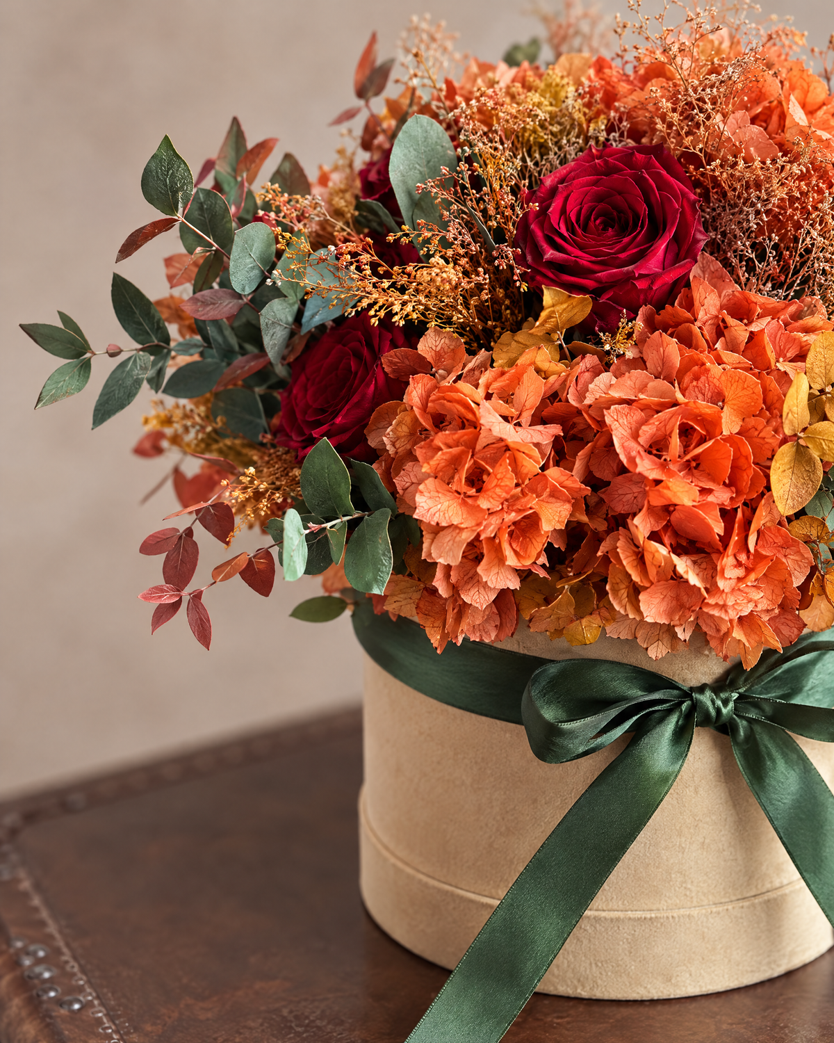 Primo piano di Velluto d'Autunno: eucalipto, ortensie arancio, rose rosse e fiocco in raso verde bosco sulla cappelliera sabbia.