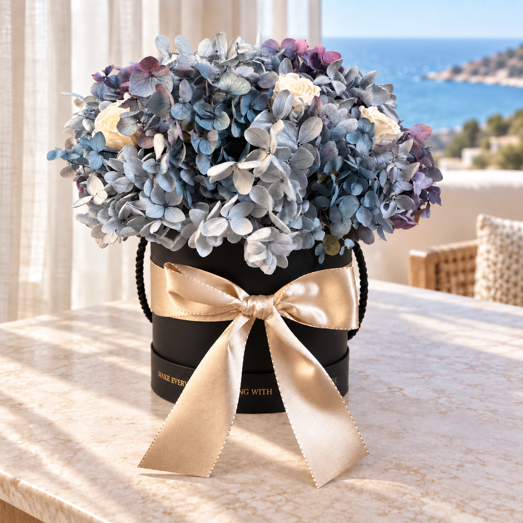 Brezza Marina su un tavolo in marmo con vista sul mare: fiori stabilizzati azzurri in cappelliera nera con fiocco champagne.