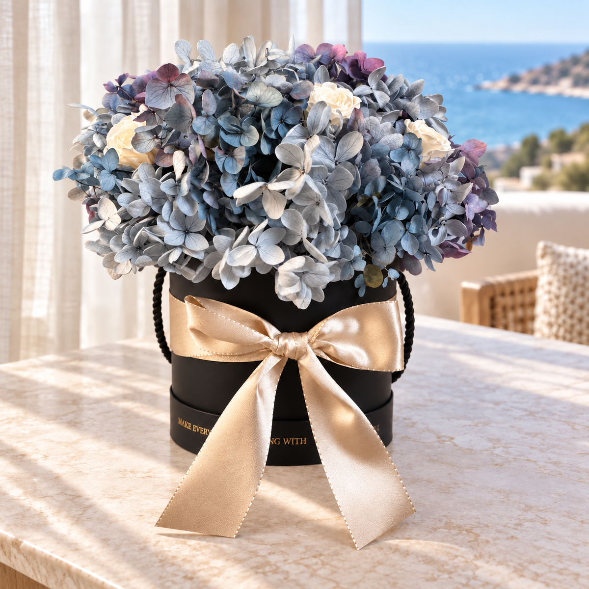 Brezza Marina su un tavolo in marmo con vista sul mare: fiori stabilizzati azzurri in cappelliera nera con fiocco champagne.
