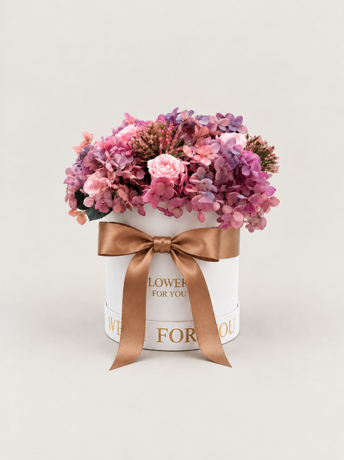 Nuvola di Seta: ortensie malva e lavanda con rose rosa cipria stabilizzate in cappelliera bianca con fiocco satinato caramello.